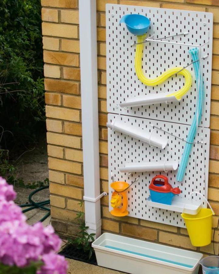 Colorful outdoor water wall station from Inspire My Play. Features tubes, buckets, and shelves for interactive summer play.