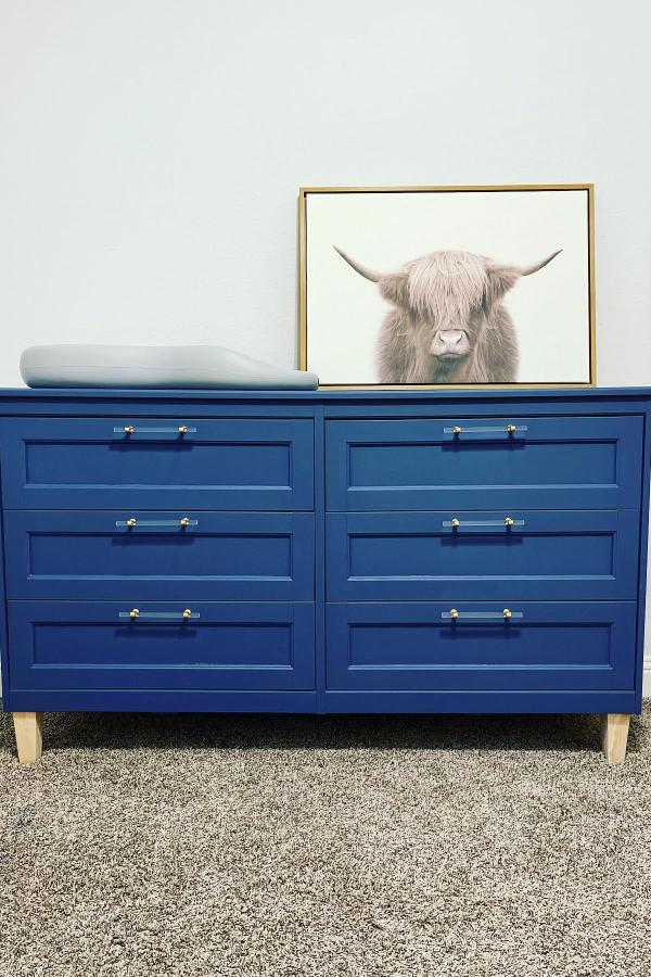 Navy painted dresser from @maddiewinchesterhome. Features brass hardware and natural wood legs on textured rug.