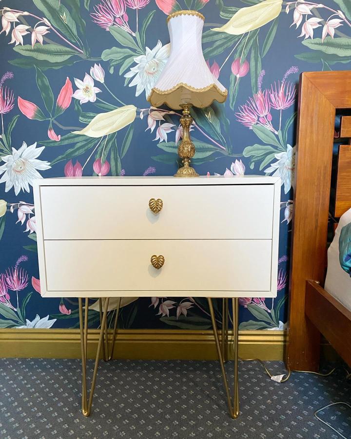White bedside table with golden hairpin legs and brass handles from @sugarstclaire. Tropical wallpaper backdrop adds colorful contrast.