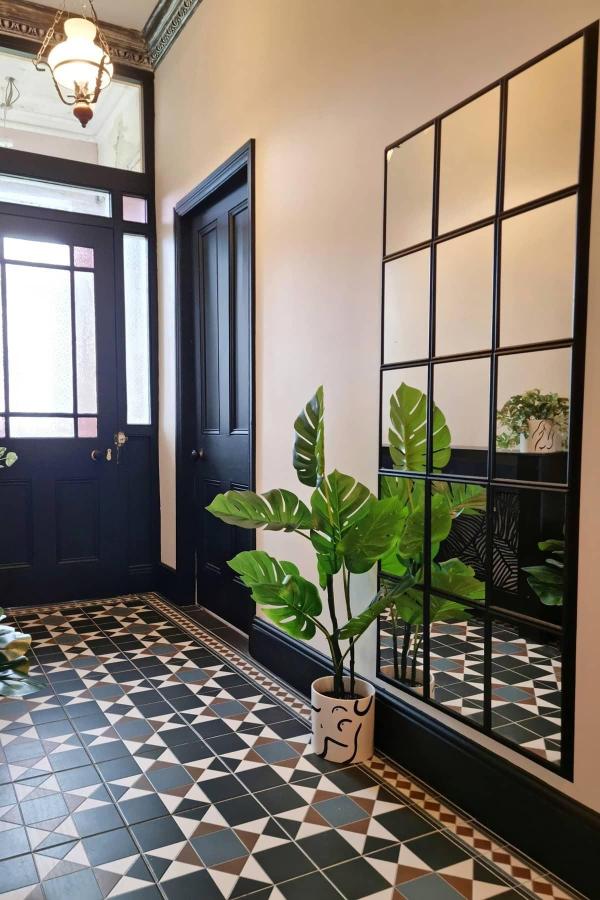 Large black grid mirror installation reflecting geometric floor tiles and tropical plant in elegant hallway entrance.