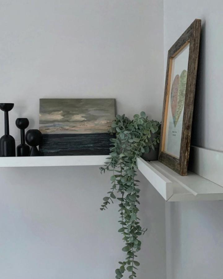 White floating shelf styled with layered artwork and trailing eucalyptus plant from @thathomelife_. Minimalist wall display setup.