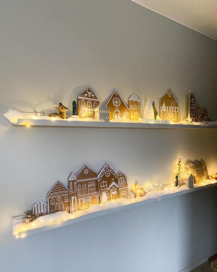 Illuminated paper house village displayed on white floating shelves from @nathaliefli. Warm string lights create magical holiday atmosphere.