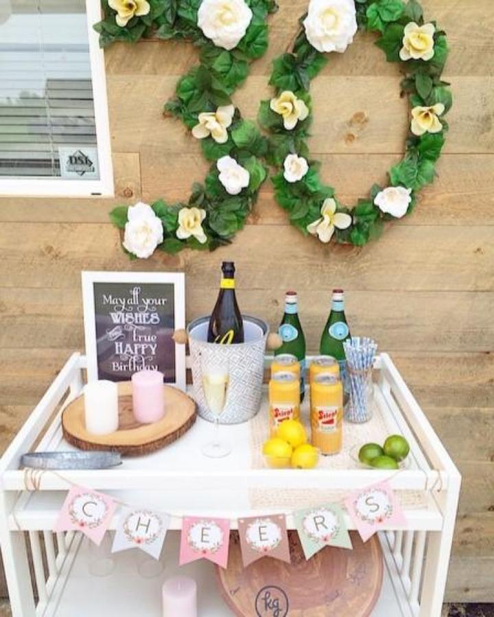 White beverage cart from @randilpearson with floral garland backdrop and pink cheers banner.