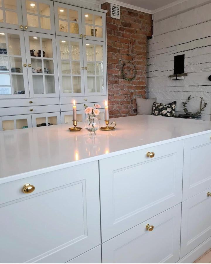 White kitchen showcasing ikea kitchen island ideas from @solsorten_paa_longisland. Glass cabinets with brass hardware and exposed brick accent.