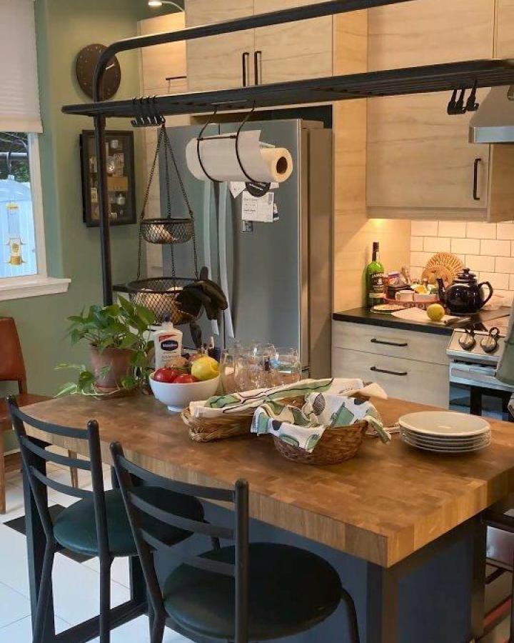 Butcher block island with overhead black metal storage rack from @inspiredkitchendesign featuring hanging baskets and kitchen tools.