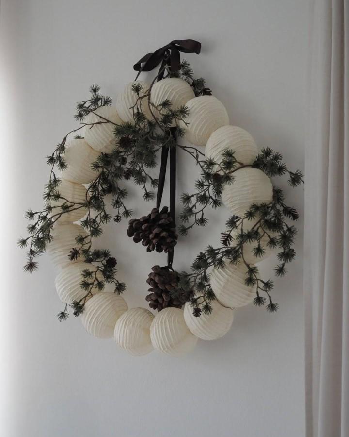 Cream paper lantern wreath from @modernity.focus. Dark pine branches and pinecones woven through accordion spheres.