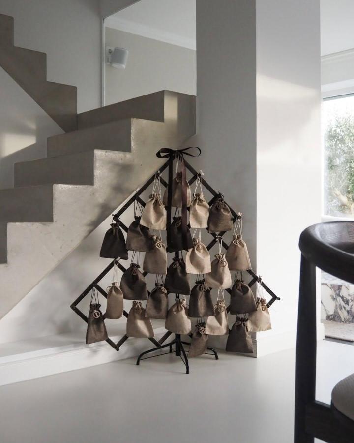 Black metal advent calendar tree from @modernity.focus with fabric pouches in neutral tones against white staircase.