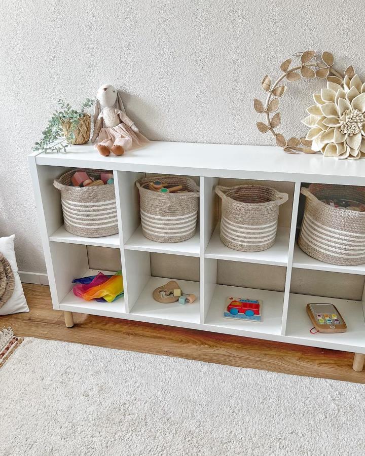 White Kallax storage unit from @dekleinemaan. Features wooden legs and striped storage baskets with toys.