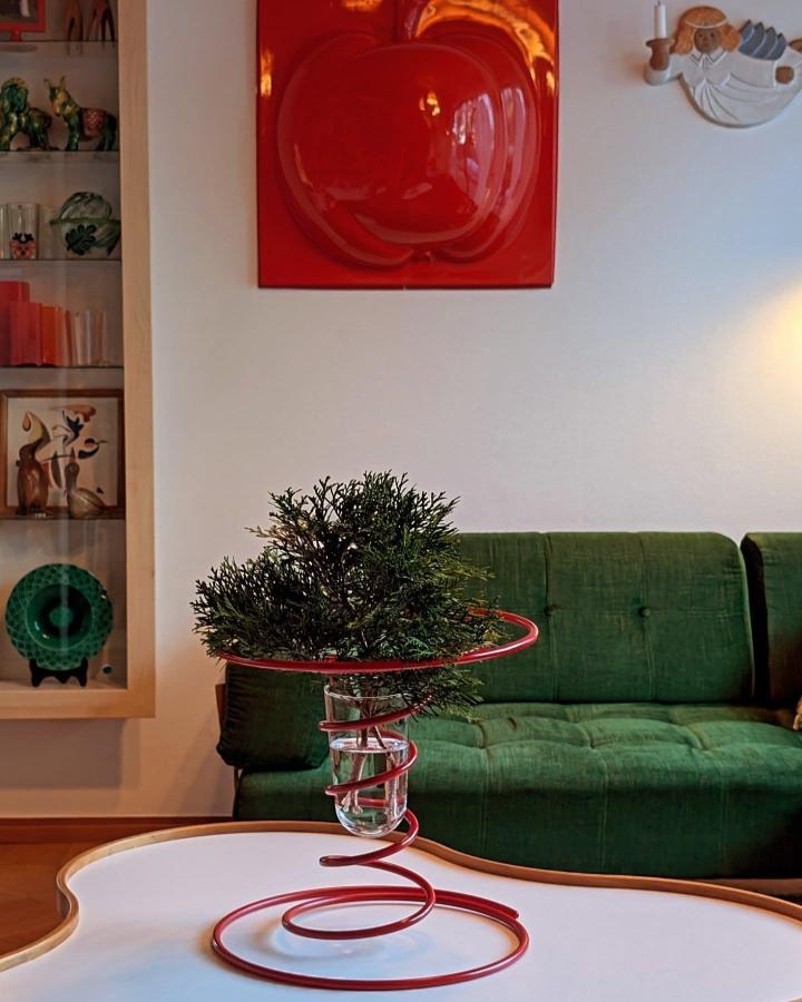 Red wire Vinterfint holder with greenery from @homebyholm. Modern centerpiece on white table beside green sofa.