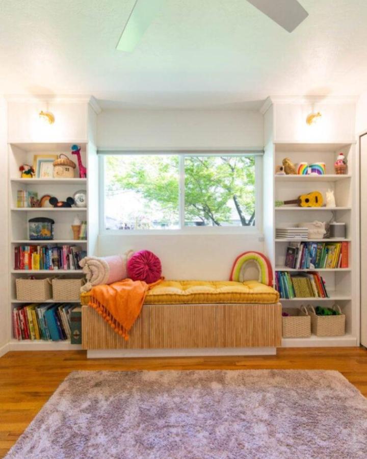 White built-in window bench with bamboo facade from Lovely Indeed featuring flanking bookshelves and colorful cushions.