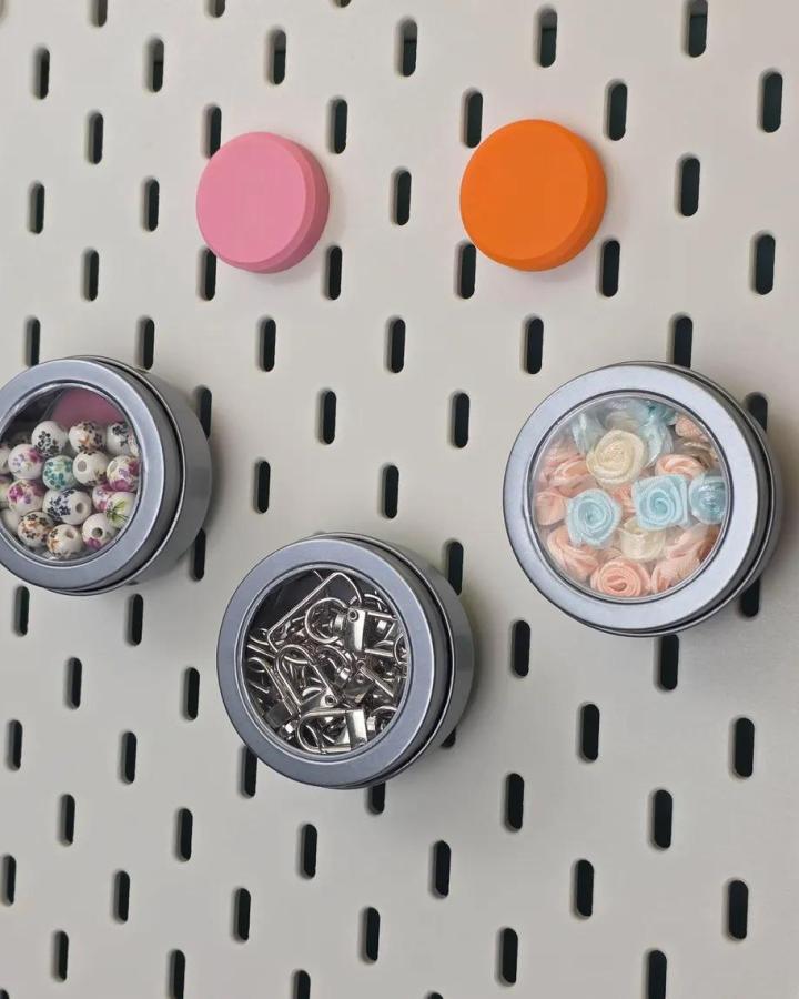 Pink and orange magnetic knobs and small storage tins on white ikea skadis pegboard craft room hacks from NookingtonsCo. Magnetic storage for craft supplies.