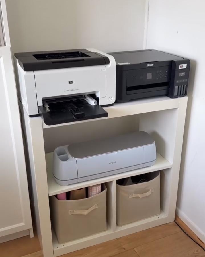 White Ikea Kallax shelf with beige storage bins and printers holding cricut tools from @hercrafts.uk. Neat and minimal ikea cricut storage ideas.