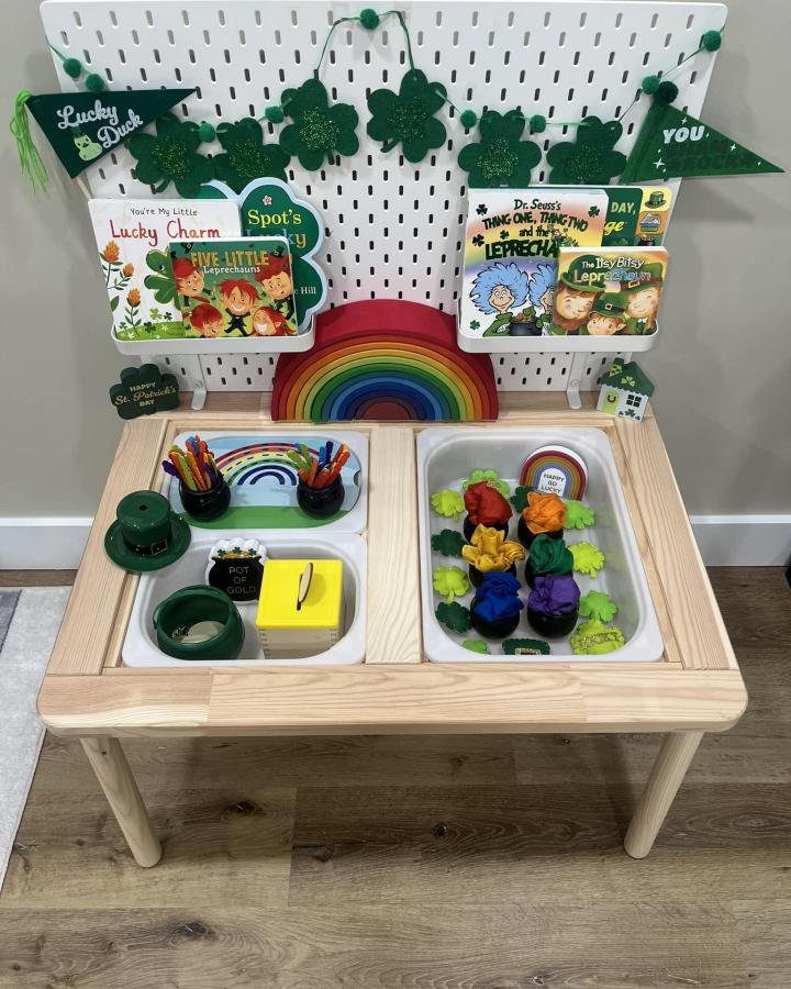 Green and natural wood Ikea Flisat table with St Patrick’s Day decorations and craft supplies from Mini Locke. Festive rainbow and shamrock elements.