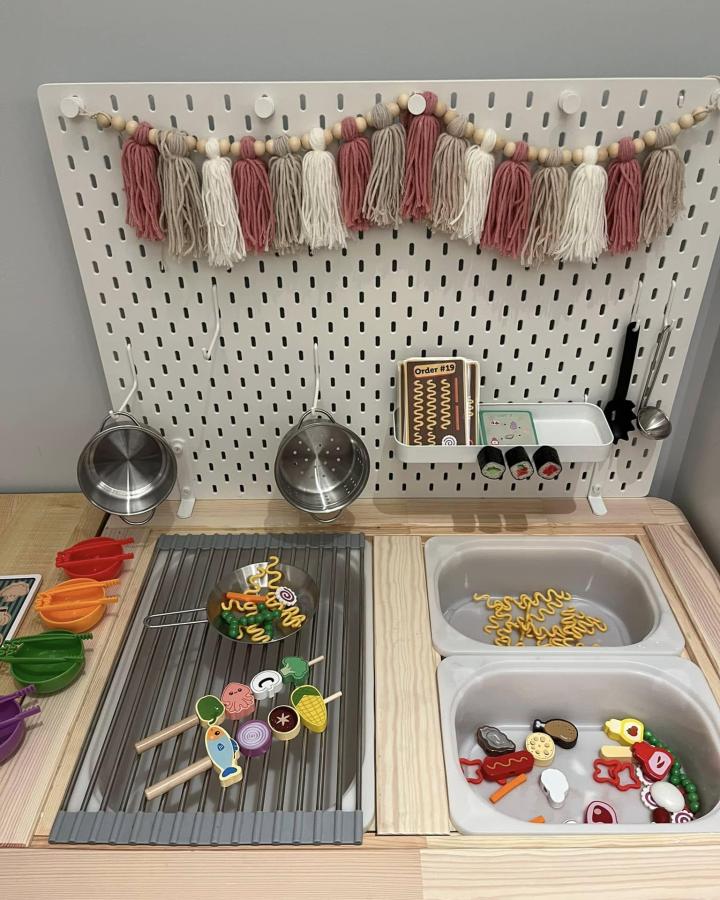 Tassel and bead decorated pegboard play kitchen on natural wood Ikea Flisat table hacks & ideas from Teale Orrick-Rudzki. Colorful toy food and cookware included.