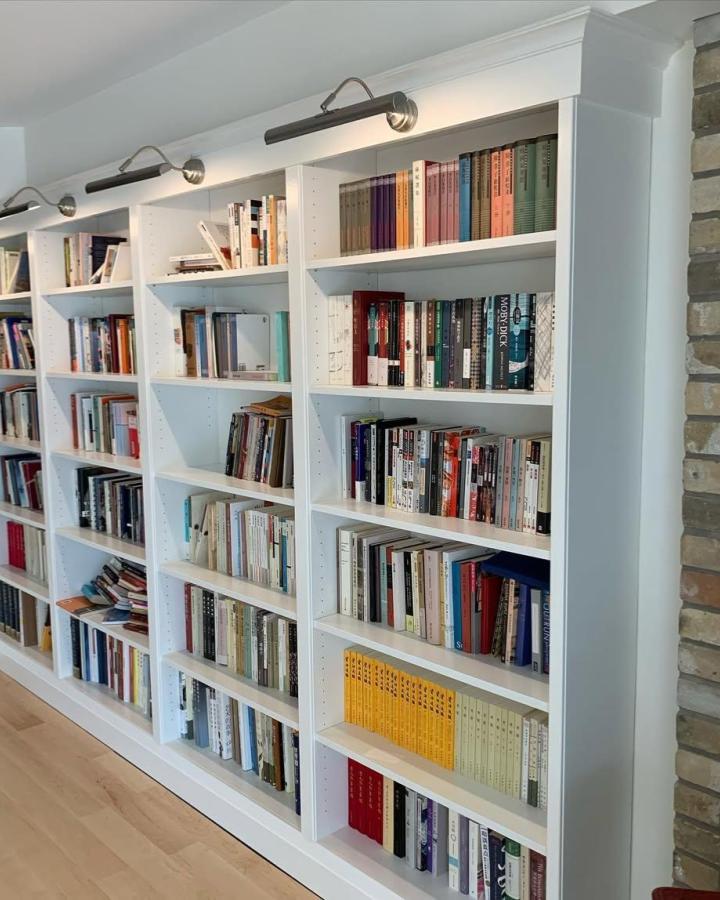 White built-in ikea billy library wall hacks in a bright room featuring crown molding and modern picture lights from @rachelpoulterinteriors. Classic, clean, and functional library wall.