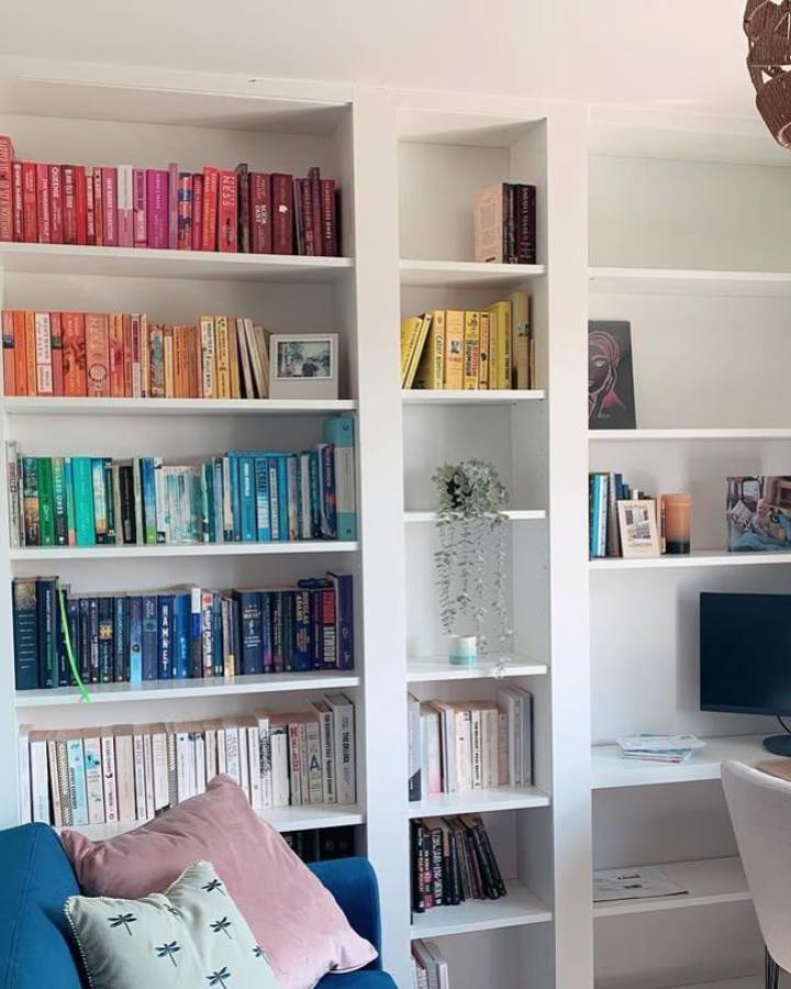 White Ikea Billy library shelves styled by @smallbookreviewsbyjo with color-coded books and green plant accents in cozy living space.