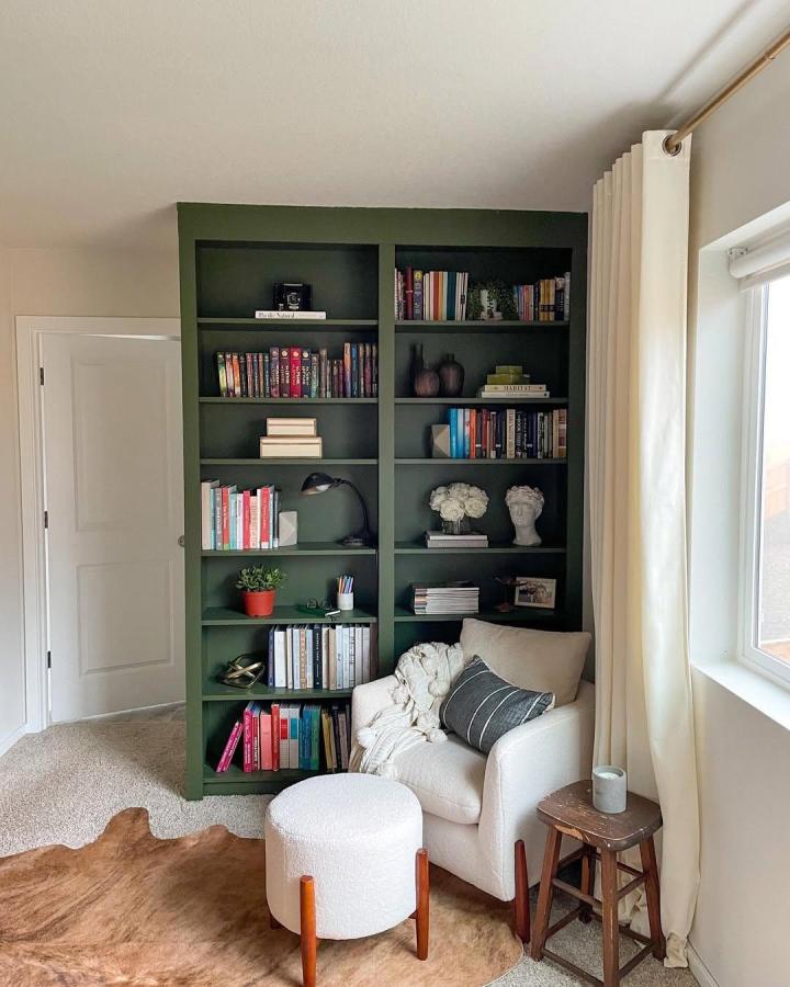 Dark olive green painted Ikea Billy library wall with books and decor by @abigailamirahome cozy reading nook with pouf and chair next to window