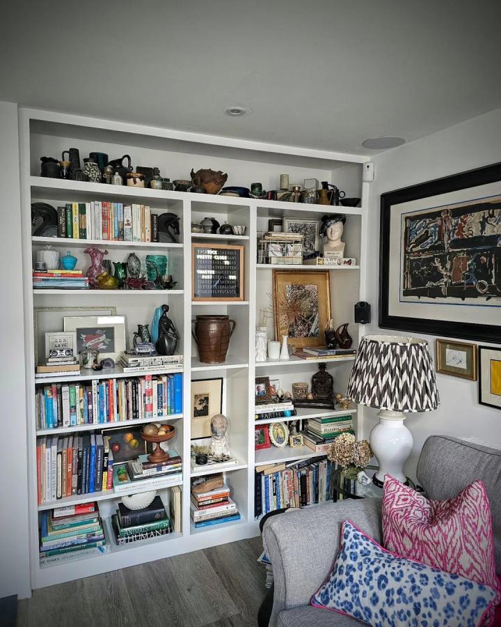 White modular ikea billy library wall hacks with mixed sized shelves filled with books, art pieces, and decor from @the_home_curator_. Bright cozy living room corner with patterned pillows and lamp.