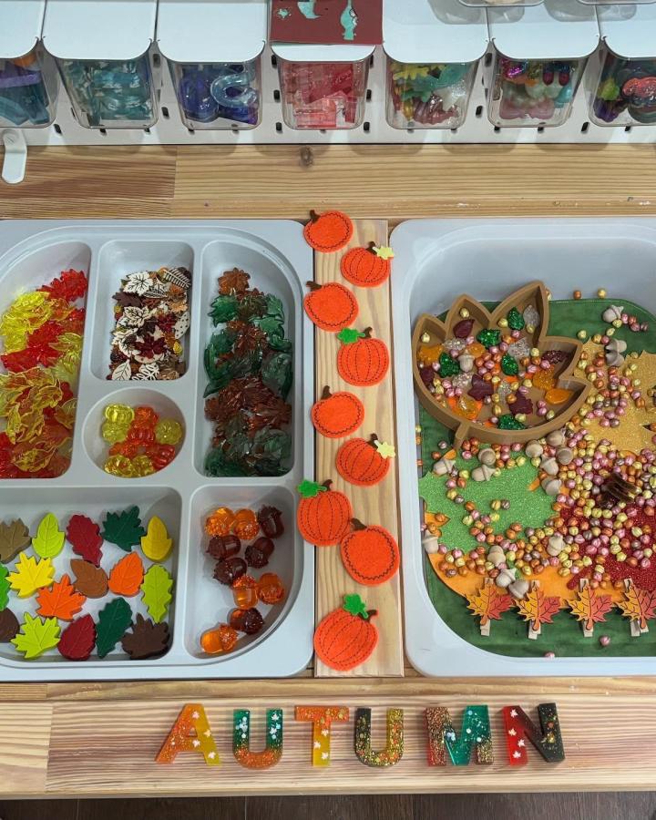 Orange and brown autumn-themed sensory table trays with plastic leaves and pumpkins from @jenniedancer81. Divided trays with colorful seasonal play items and wooden pumpkin decorations.