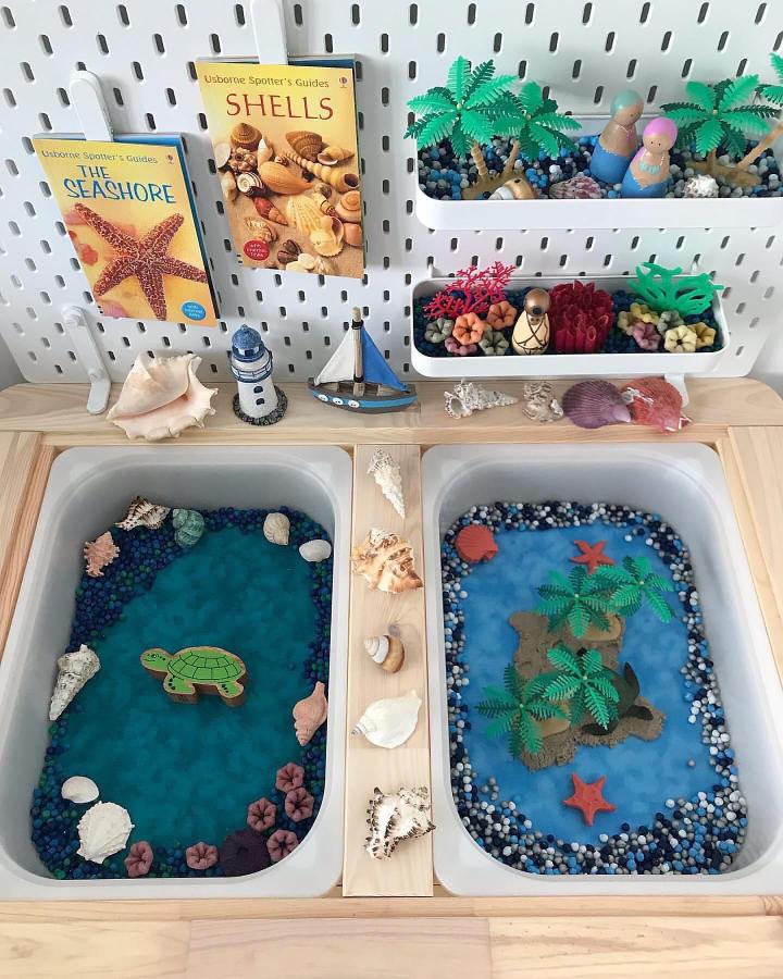 Beachy Ikea Flisat sensory table ideas with blue beads, shells, mini palm trees, and sea creature figures from @theplaymatkingdom. Coastal sensory play setup.