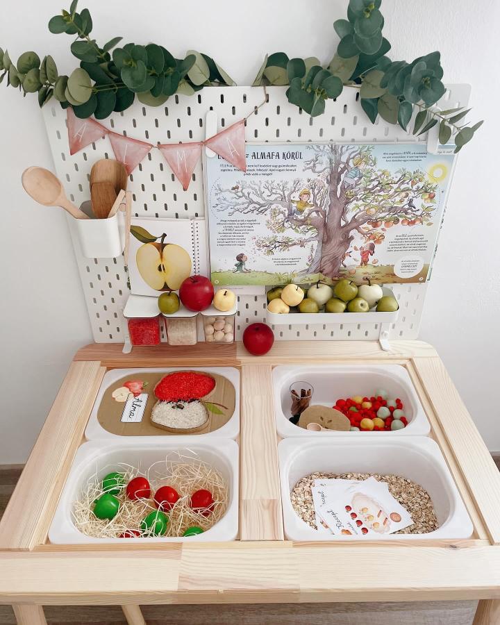 Natural pine wood sensory table with white bins and pegboard storage from @panna_asztalkaja. Includes colorful fruits and tactile materials ikea flisat sensory table ideas.