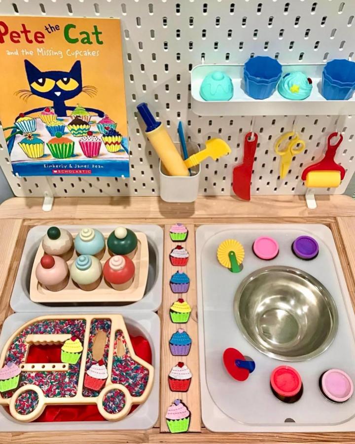 Colorful wooden cupcake sensory play station on Ikea Flisat table by @playing_the_day_away with pegboard utensils and mixing bowl setup.