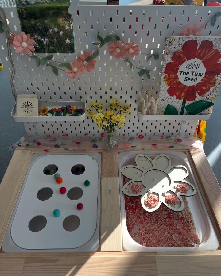 [wooden and white Ikea Flisat sensory table ideas with floral decorations and tactile play trays from @ventureintoeyfs. Sensory sorting and texture exploration setup with natural accents]