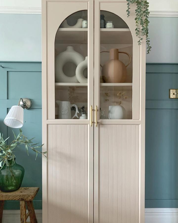 Beige painted bookcase with arched glass doors and ribbed panels from @crack_the_shutters. Styled with ceramics and subtle gold handles in cozy room.