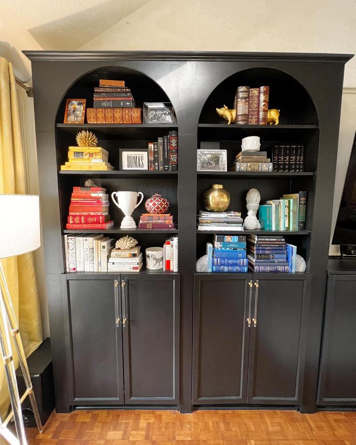 Black matte archway bookcases with color-organized books and gold decor from @pinchmeimhome stylish storage and display