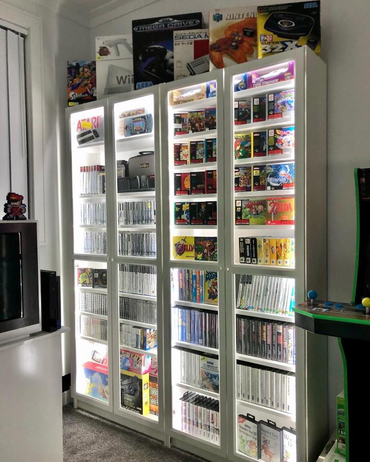 White Ikea Billy bookcases with glass doors holding colorful vintage video game cases illuminated by LED strips for a retro gaming collection display.