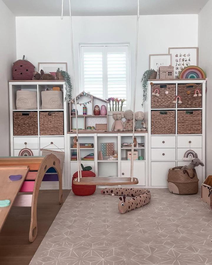 Natural wicker and white Kallax shelves with pastel toys and green plants in a child’s playroom setup by @latteandlullabies. Cozy, organized toy storage.