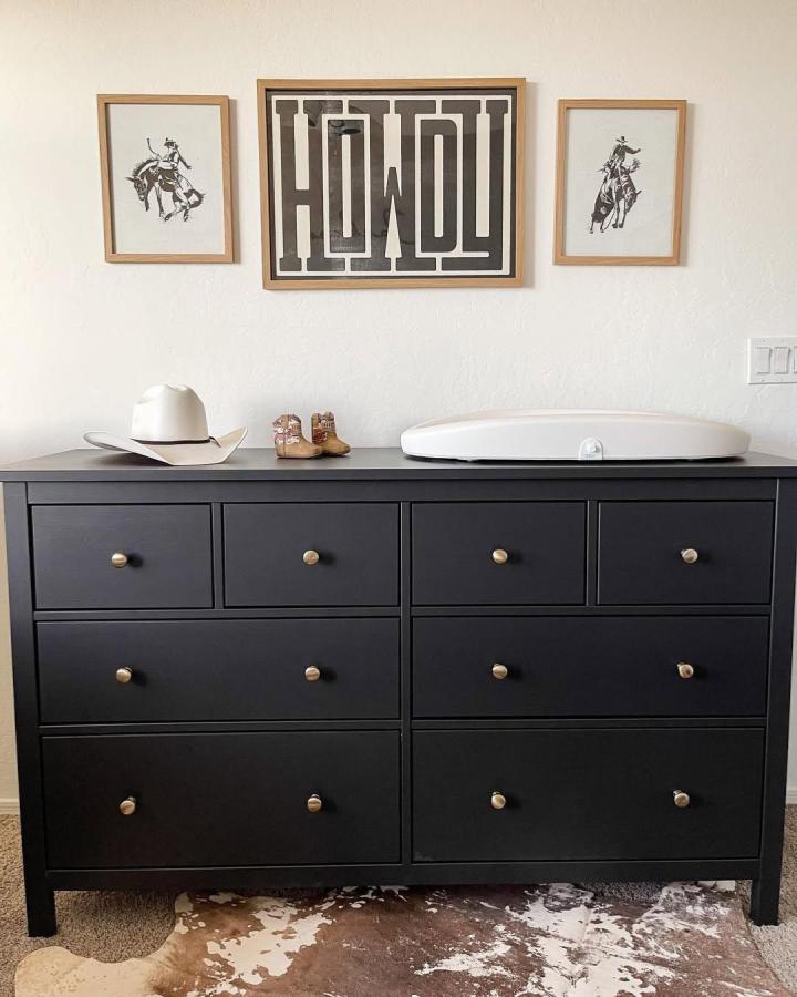 Black IKEA Hemnes dresser with brass knobs and white changing pad styled with cowboy boots and Western art for ikea nursery dresser ideas.