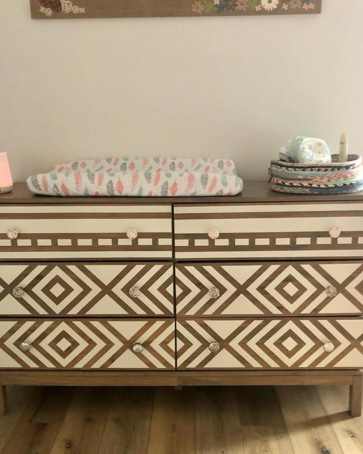 Warm wood and white geometric patterned nursery dresser with decorative knobs from @blackburn.custom.homes. Eleven drawers with diamond and square designs.