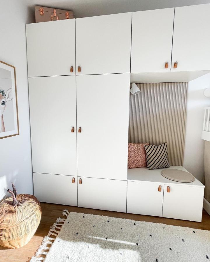 White modular storage with cozy nook and leather handles from @anetas_leben. Functional kids room setup with textured wall panel and cushions.