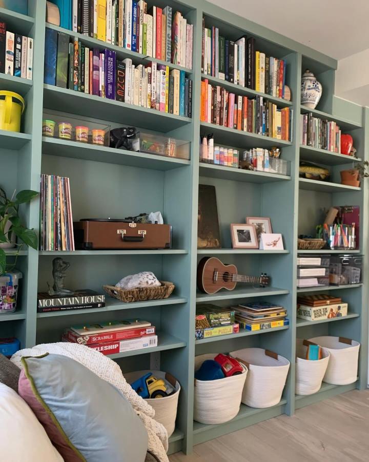 Soft green painted built in shelving unit from @griffiths_kirsty with books, baskets, and games creating a cozy organized space.