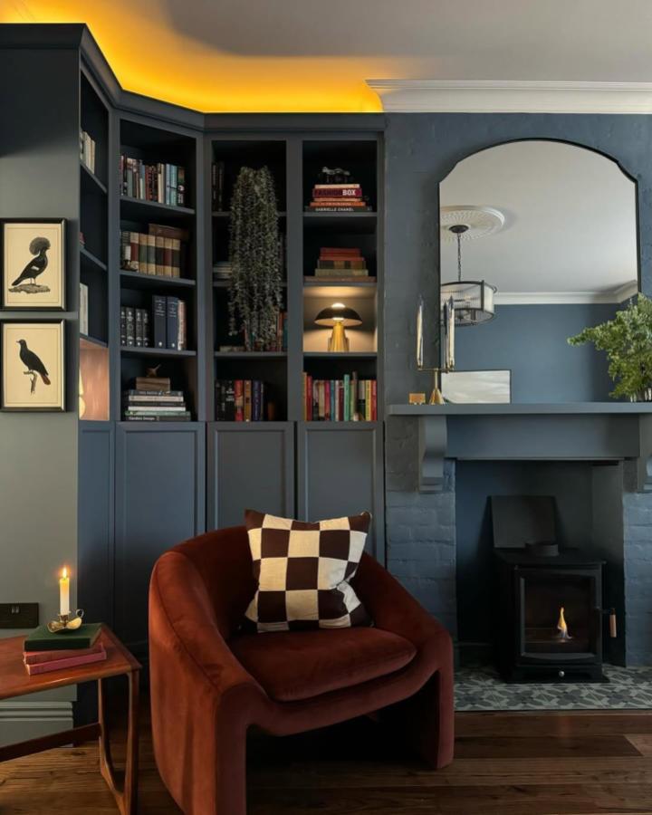 Dark blue painted built in with warm LED lighting and cozy chair from @fantasyandfoliage. Moody reading corner with brass and greenery.