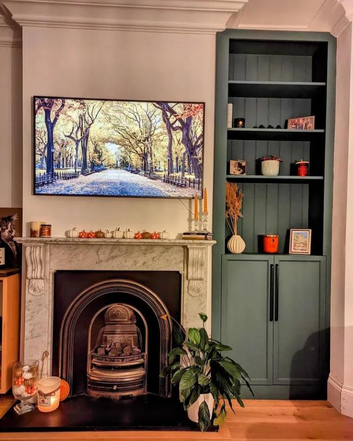 Green painted built-in bookshelf with vertical paneling and black handles next to fireplace from @cc_woodwork. Cozy media wall design with shelves and cabinets.