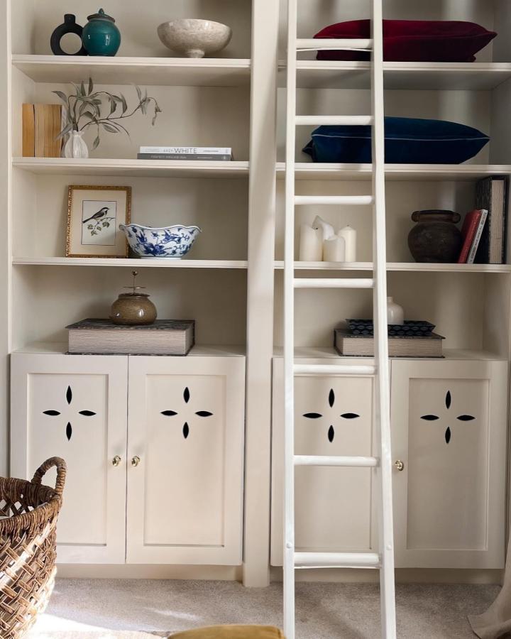 White painted wooden bookcase with decorative cabinet doors and a ladder from @painted_by_alexa elegant room setup with shelves and cushions.