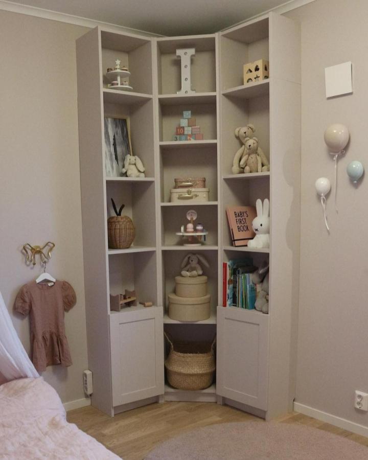 Beige corner bookshelf with mixed open and closed storage by @kuttergatan in a child's pastel-themed room with soft toys and wicker accents.