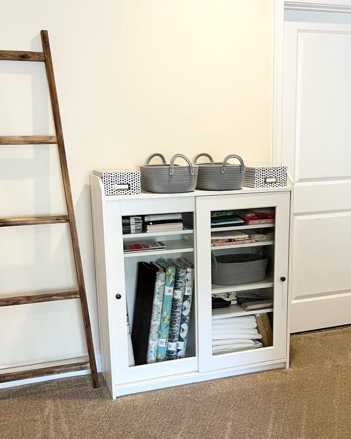 White Ikea Hauga cabinet with glass doors and grey woven baskets in a craft room by @sewthurstyquiltco. Organized fabric storage and rustic ladder detail.