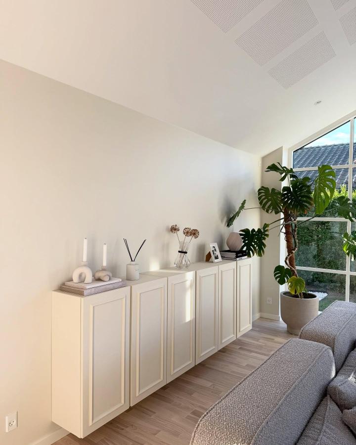 Cream white minimalist wall-mounted storage unit from @yawzzi. Smooth panel doors with elegant classic design near cozy living room window.