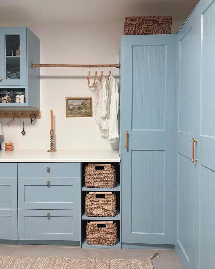 Soft blue cabinetry laundry space by @ccloudyhills with woven basket shelves and brass accents creating cozy storage vibes.