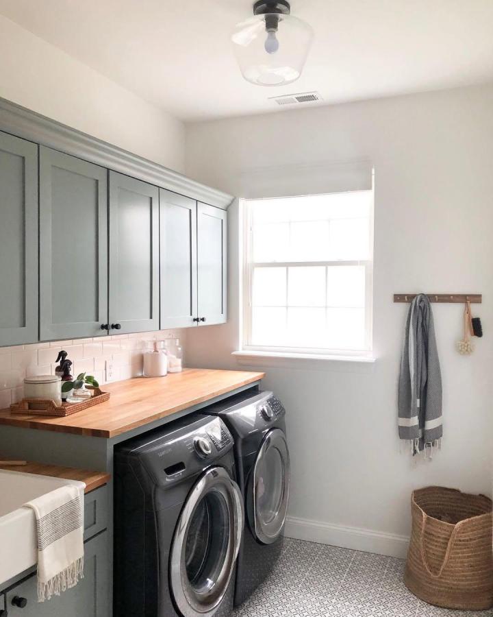 Muted green cabinetry with butcher block countertop Ikea laundry room storage from @theeveryhome featuring apron-front sink and front-load washer dryer.