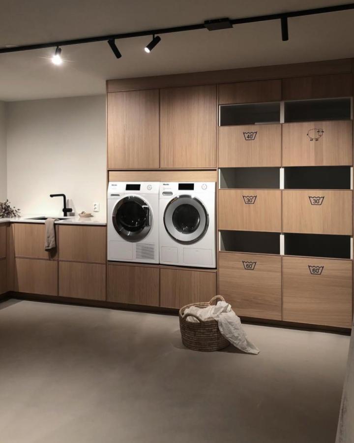 Wood-toned laundry cabinetry from @byschedin featuring built-in washer-dryer and labeled open shelves for sorting.