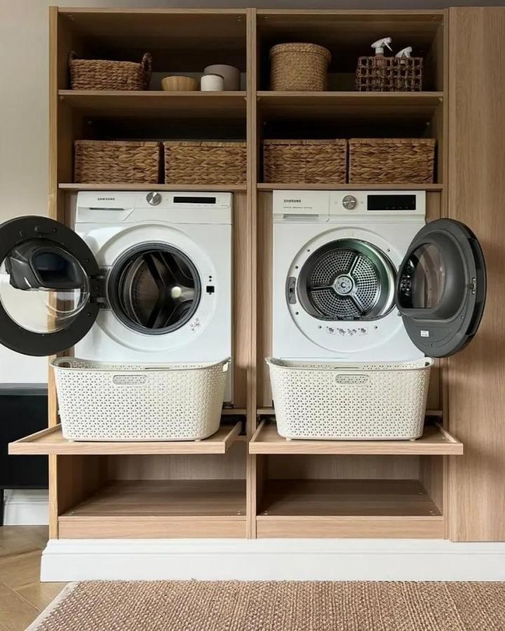 Soft oak finish laundry space from @klk.interiors featuring pull-out shelves, woven baskets, and organized washer dryer setup.