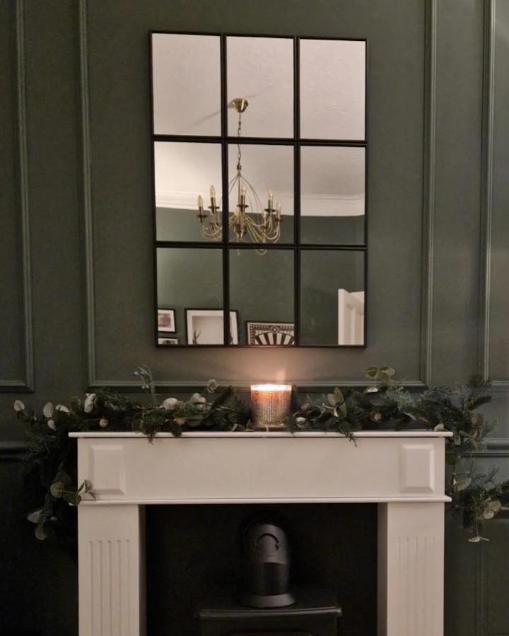 Dark framed ikea mirror wall on deep green wall above white fireplace by @our_little_sandstone reflecting brass chandelier in cozy room.