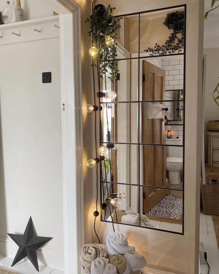 Warm black grid panel mirror with string lights and soft towels by @flowbylara reflecting cozy home entryway and bathroom details.