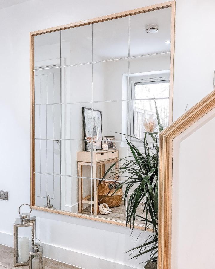 Bright grid mirror wall with natural wood frame by @homewithlucie reflecting a cozy, airy interior space with plants and soft lighting.