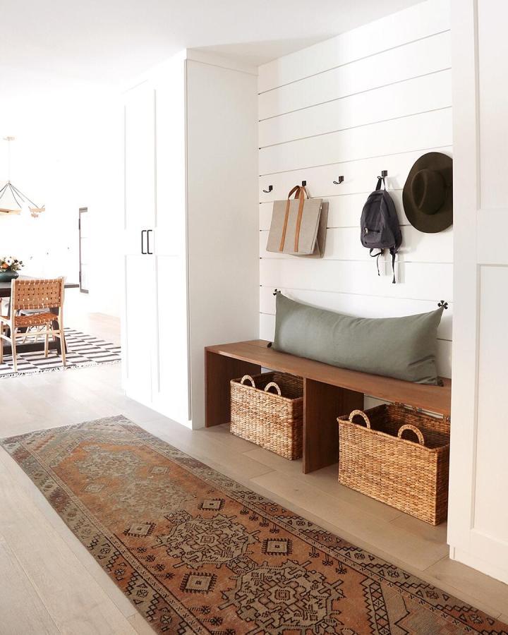 White and wood compact mudroom with built-in storage and woven baskets by @jennykomenda. Cozy bench with sage cushion and black hooks.
