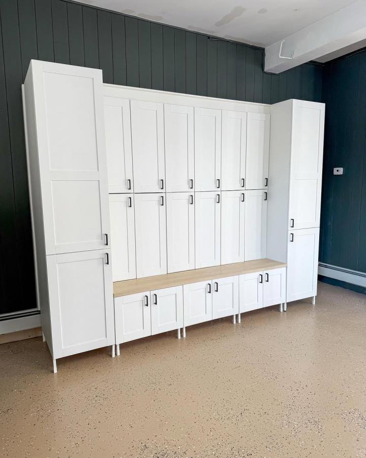 White Shaker style mudroom locker storage with wood bench and black handles in front of dark green walls by @reno.by.rach. Sleek modular storage solution.